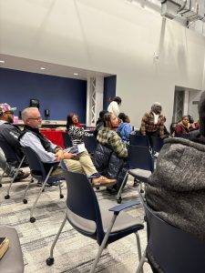 People sit and stand in a brightly lit room with blue walls, attending what appears to be a meeting or public event.