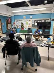 Three people sit at tables in a community room, engaged in discussion near a bulletin board and a Christmas tree.