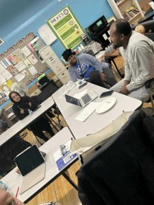 Four adults sit around tables in a classroom, talking and smiling; laptops and papers are on the tables.