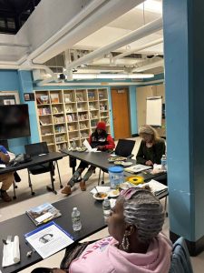 Four people sit around tables in a classroom with bookshelves, papers, food, and drinks visible on the tables.