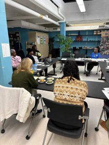 Six people sit around tables in a meeting room with food plates, documents, and a large plant in the background.