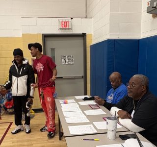 Two young men stand talking near a table where two older men sit reviewing paperwork in a gymnasium.