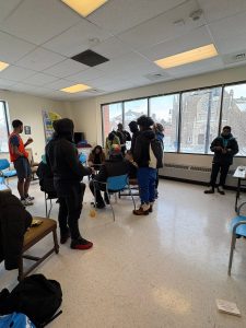 A group of people gather and talk in a classroom with large windows showing a snowy scene outside.
