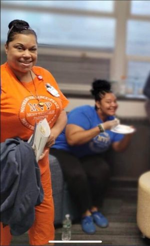 Two women smile indoors; one stands holding papers, the other sits eating from a plate. Both wear shirts with the same logo.