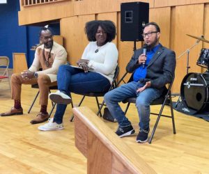 Three people sit on chairs in a gymnasium; one person speaks into a microphone while the others listen.