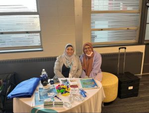 Two women sit behind an information table with brochures, pens, and giveaway items in a brightly lit room.