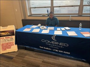 A woman sits behind a table with insurance brochures and a phone, next to a sign advertising various insurance types.