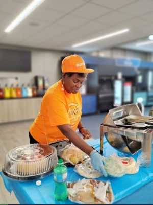 Woman in orange shirt and cap prepares food at a counter with bottled drinks and desserts in the background.