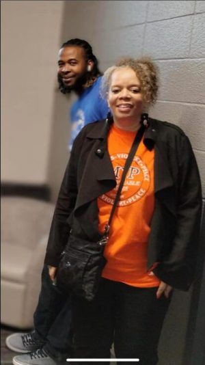 Two people stand indoors by a wall; one wears an orange shirt and black jacket, the other stands slightly behind.
