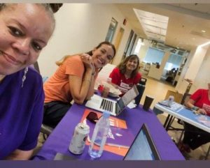 Four women sit at a table with laptops, water bottles, and papers in a brightly lit office setting.