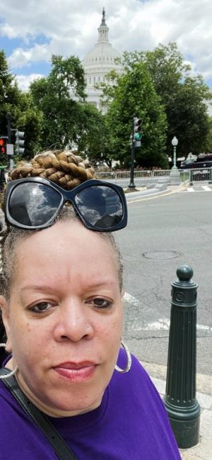 Woman with braided hair and sunglasses poses on a street with the U.S. Capitol building visible in the background.