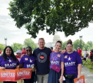 Five people stand outside near the Capitol, some wearing “Brady” shirts and holding “United Against Gun Violence” signs.