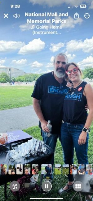 Two people stand together outdoors by a table with wrapped food, both wearing “Enough” shirts at a park event.