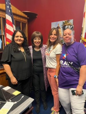 Four women stand together in an office with an American flag and a desk visible in the background.