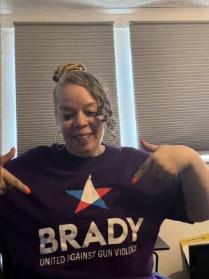Woman pointing to her “BRADY United Against Gun Violence” shirt while sitting indoors in front of closed window blinds.