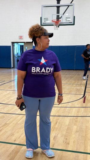 Woman in a purple “Brady: United Against Gun Violence” shirt stands on an indoor basketball court holding a phone.